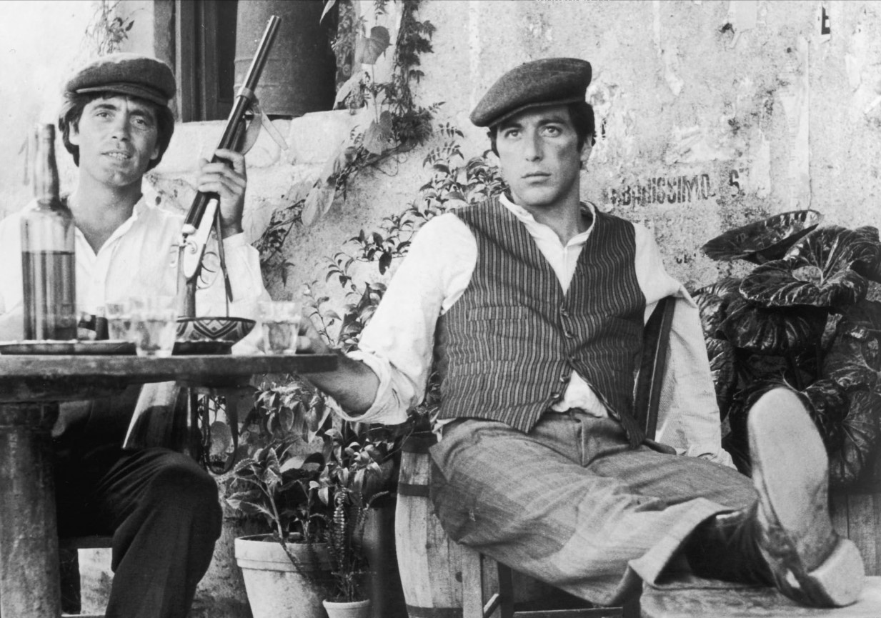 michael corleone and somebody else taking a seat near a table in their home village